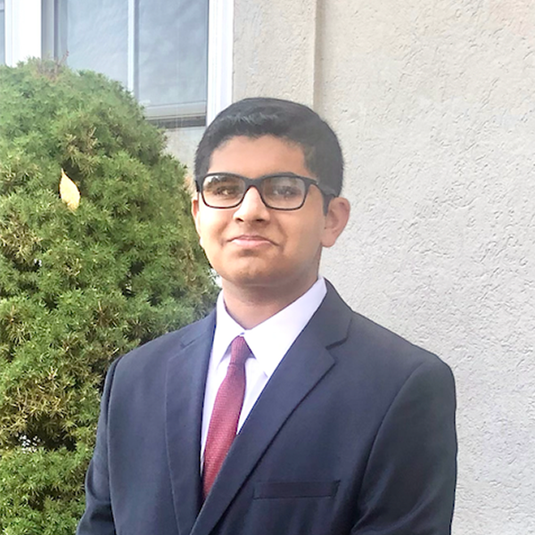 Young man in glasses and a dark suit with red tie poses outdoors beside a shrub and building.