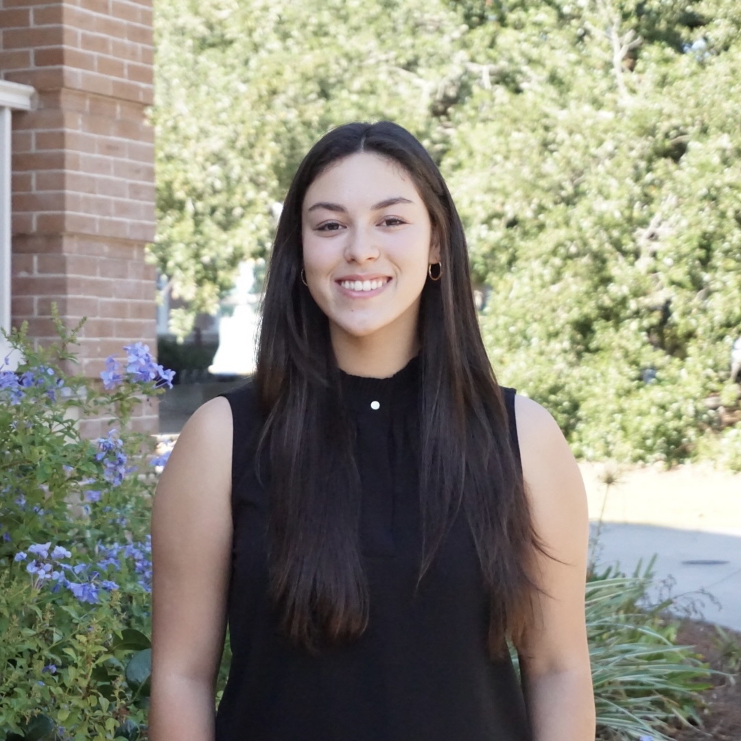 Smiling young woman with long dark hair in a black top, outdoors.