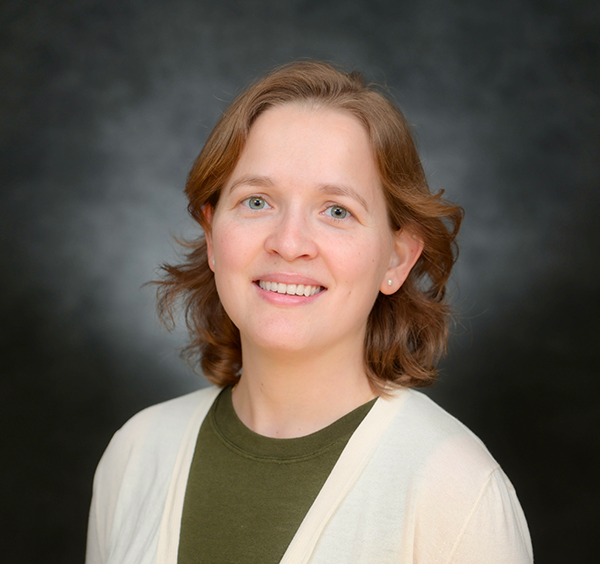 Smiling woman with light brown hair, green shirt and cream cardigan against a dark grey background.