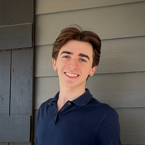 Smiling young man with brown hair and blue eyes poses in a navy polo shirt against gray wooden siding.