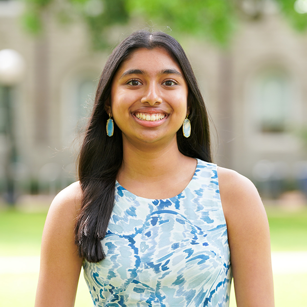 Smiling young woman with long dark hair and blue earrings poses outdoors in a blue-and-white patterned sleeveless top.