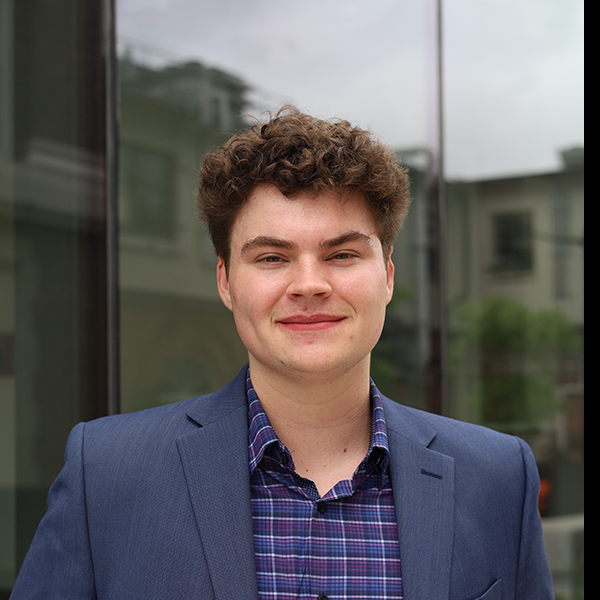 Smiling young man with curly brown hair wears a blue blazer and purple plaid shirt, posing outdoors near a modern glass building.