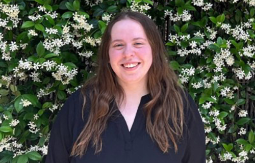 Smiling young woman with long brown hair in a black top, against white flowering vines.