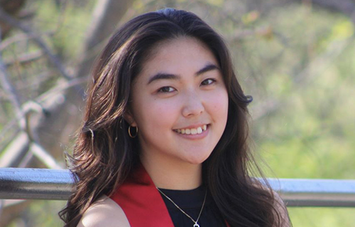 Young person smiling, wearing a black top and red sash in an outdoor setting