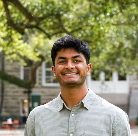 Young man with dark hair smiles wearing a light green shirt outdoors.